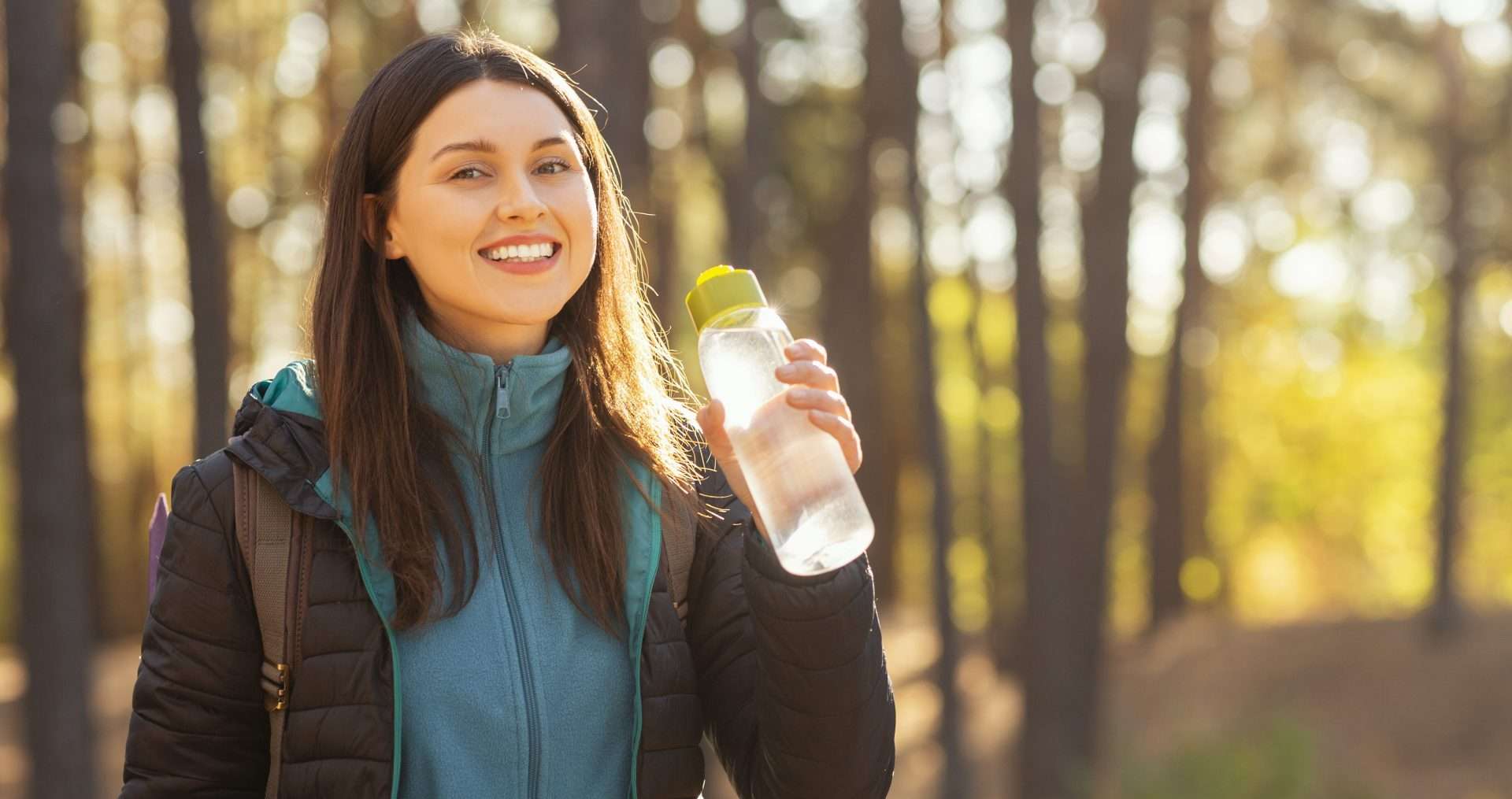 Cheerful young woman drinking water while hiking in autumn forest, panorama with empty space