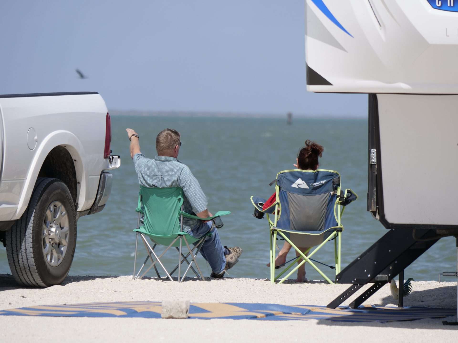 Two people sitting in front of RV on outdoor rug on the beach with lots of sand.