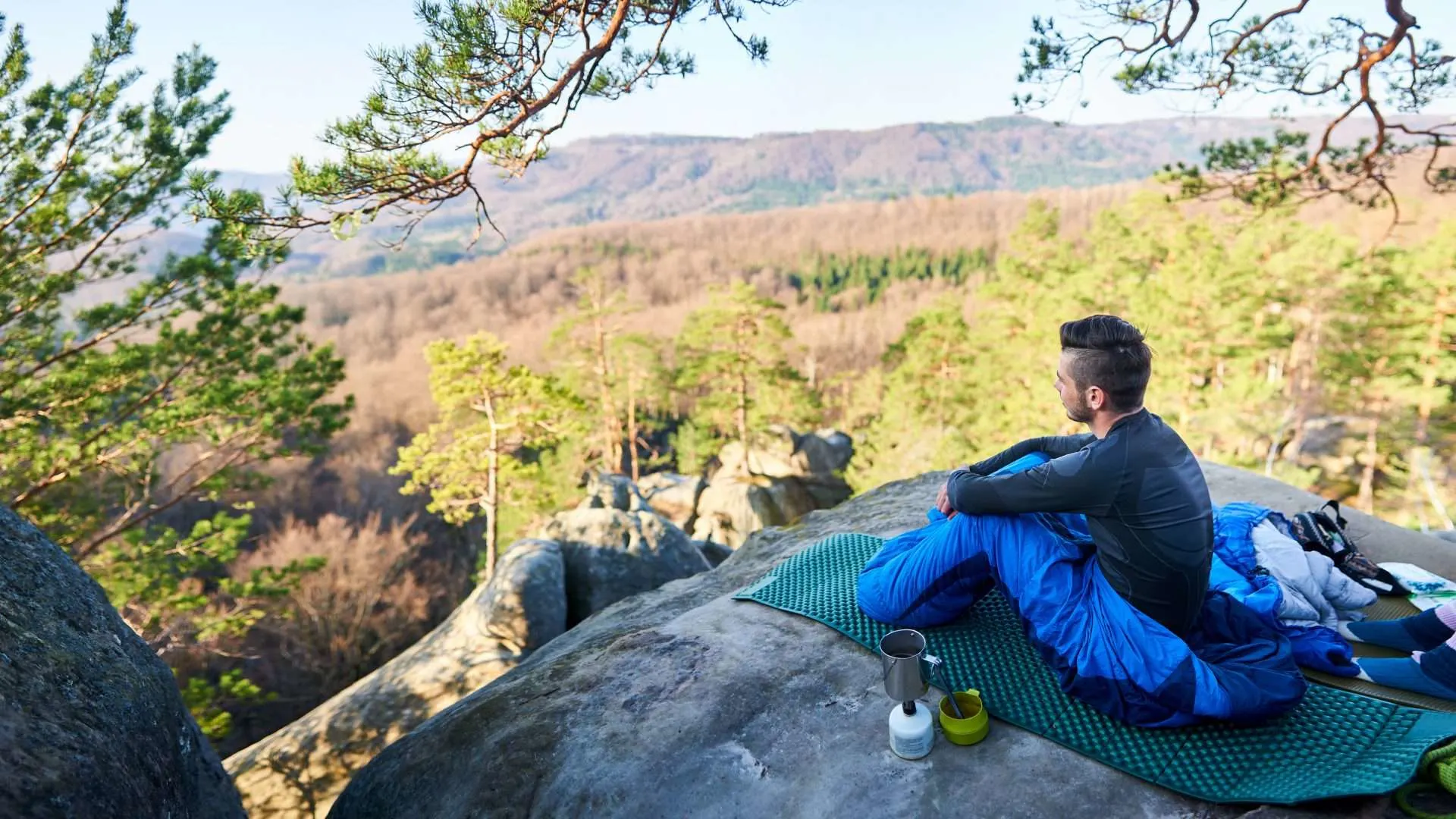 Man sitting inside bivy sack on a cliff looking out at forest