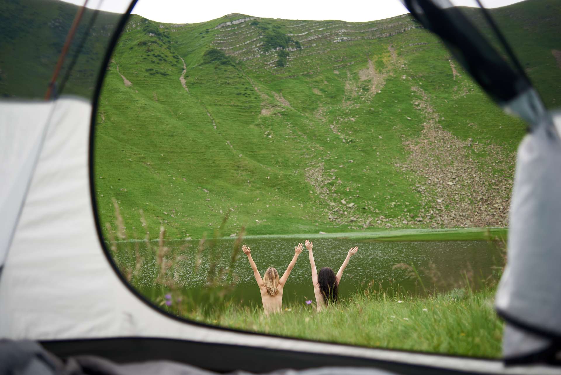 Back view of two naked female travellers lifting hands up in the air near the lake in the mountains. View from tent inside. Lifestyle concept adventure summer vacations outdoor