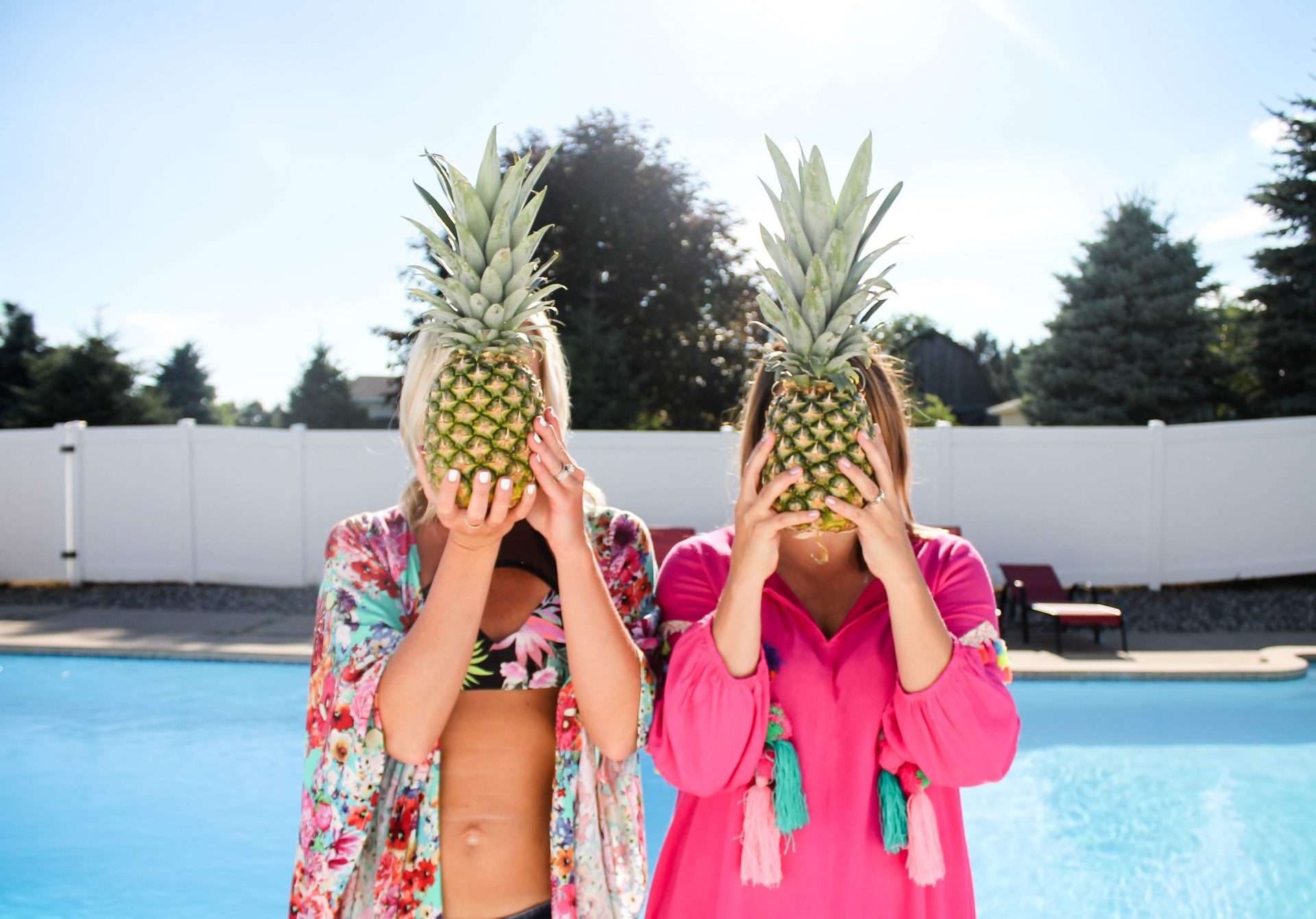 Two women holding pineapples in front of their faces by pool